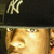 A person wearing a black cap with white NY logo, showing a side profile with visible ear and nose, under warm lighting.