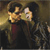 Two figures in dark clothing stand side-by-side against a blurred background with warm yellow and brown tones.