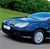 A blue sedan with silver accents appears in profile view on a gray road, set against a green grassy hill and dark green foliage background.