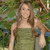 A pixelated image of a woman with long brown hair wearing a sleeveless green dress, standing outdoors with green foliage in the background.