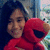 A person with long dark hair holds a red plush Elmo toy against a blurred background with light-colored vertical elements.