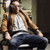 Pixelated figure in brown jacket and white shirt seated with legs crossed, holding a cigarette, against muted background.