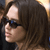 Pixelated profile view of a person with long dark hair wearing dark sunglasses and a dark top against a blurred background with light and dark tones.