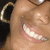 A person with dark skin and large hoop earrings smiles broadly, showing teeth, in a close-up view with a dark background.