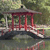 Static pixelated image of a red pagoda-like structure with dark roof and columns, situated on a dark platform over water, surrounded by green foliage.