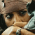 Pixelated close-up of a figure with light brown skin, dark eyes, and a brown head covering, resting a hand near the chin against a dark background.