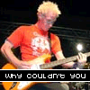 A person with short blonde hair wearing an orange shirt plays a light brown electric guitar on a dark stage with stage lights visible.