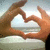 Two hands form a heart shape against a light blue sky background with faint clouds.