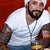 A person in a white t-shirt and turban holds a yellow object against a red background, with visible tattoos on arms.