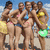 Group of women in swimwear posing on a beach with yellow and green props under blue sky.