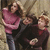Three people stand closely together in a pixelated image; the person on the left wears a pink top and dark pants, while the other two wear dark clothing with red accents.