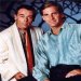 Two men in formal attire sitting side-by-side with a dark background, rendered as a low-resolution pixelated image.