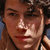 A pixelated image of a person with dark curly hair, light skin, and a neutral expression, cropped to show the upper chest and shoulders against a blue background.