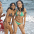 Two women in bikinis stand on a beach with ocean waves behind them; one woman wears a green bikini, the other a dark bikini.