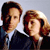 Two people in formal attire stand side-by-side against a light gray background, one with dark hair and the other with light brown hair.