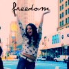 A person with long dark hair raises both arms above their head, holding a large handwritten-style 'freedom' text against a blurred cityscape background.