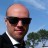 Pixelated portrait of a bald man wearing dark sunglasses and a black suit with white shirt collar visible against blue sky background.