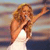 A woman with long wavy brown hair sings into a microphone, wearing a white sleeveless dress against a blurred background of horizontal color bands.