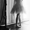 Black-and-white image of a person in a short dress standing near a microphone stand with a bright light source behind them.