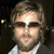 Pixelated close-up of a person with light brown hair, wearing dark sunglasses and a light-colored collared shirt against a dark background.