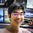 A pixelated static image shows a person with dark hair and glasses, wearing a light-colored shirt, against a background with blue and white elements.