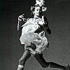 Black-and-white pixelated image of a person in a dynamic pose holding a microphone, wearing a patterned dress and dark tights.