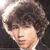 Pixelated close-up of a person with curly brown hair and light skin, wearing a white collar against a dark background.