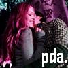 A static image shows two people in close physical proximity, one with long pink hair and the other with short dark hair, against a dark background with blurred green and purple elements.
