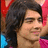 A person with long dark hair and a light brown complexion wears a pink garment over blue, set against red and white background elements.