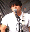 A pixelated static image shows a person with short dark hair holding a microphone near their mouth, wearing a white shirt against a blurred background.