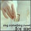 A hand holds a small rectangular object against a light background with text at the bottom reading 'sing something sweet for me'.