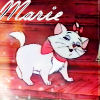 A white cartoon cat with a red bow and scarf stands on a red background beneath the text 'Marie' in white cursive.