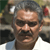 A pixelated close-up of a person with short graying hair, dark eyes, and a mustache, wearing a light-colored shirt against a blurred background.