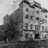 Black-and-white pixelated image of a multi-story building with rectangular windows and a sloped roof, partially obscured by dark foliage on the left.