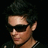 Pixelated close-up of a person with dark hair, wearing dark sunglasses and a dark shirt with a checkered pattern on the shoulder, against a black background.