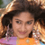 Pixelated static image of a person with curly dark hair wearing an orange top and purple garment, partially visible face with neutral expression, background includes light brown tones and blurred elements.