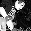 Black-and-white image of a person with short hair wearing a dark shirt with a striped pattern on the sleeve, holding a guitar-like instrument.