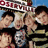 A static image shows multiple people in a group pose with a blue sky background and partially visible red and white text at the top.