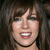 A pixelated close-up of a person with brown hair, dark eyes, and a wide smile showing teeth, wearing a black choker necklace against a dark background.