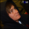 A person in a black hooded garment with a white zipper pull, positioned against a patterned background with visible text "TOM" in blue and white.