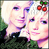 Two blonde women with blue eyes pose closely together, one wearing a white headband with red and green cherries, the other with a dark green top and black collar.