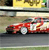 A red sports car with white accents is depicted in motion on a gray road, with a blurred background featuring yellow and green elements.