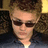 Pixelated close-up of a person with short brown hair wearing dark sunglasses and a light-colored shirt against a blurred background.