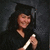 A person in a black graduation cap and gown holding a rolled diploma, smiling against a dark background.
