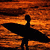 Silhouette of a person holding a surfboard against an orange sunset sky with palm trees on the right.