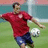 Pixelated image of a person in a red shirt and blue shorts kicking a soccer ball on a green field with a goalpost in the background.