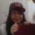 A person with long dark hair wears a dark cap with a red emblem and a dark red shirt, holding a finger near their chin against a light gray background.