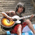 A person in red shorts and a white shirt plays an acoustic guitar against a stone wall background with blue