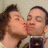Two people kiss in a mirror selfie; one holds a dark smartphone with a visible camera lens, the background is a light textured wall.