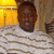 A pixelated image of a person wearing a white sweater with brown stripes, standing in front of a yellow-lit background and wooden structure.